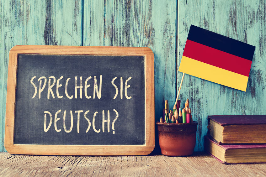 a chalkboard with the question sprechen sie deutsch? do you speak german? written in german, a pot with pencils, some books and the flag of Germany, on a wooden desk